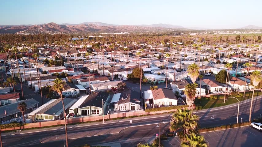 Slow drone flight capturing residential trailer park community featuring rows of small houses lined with tall palm trees at sunset.