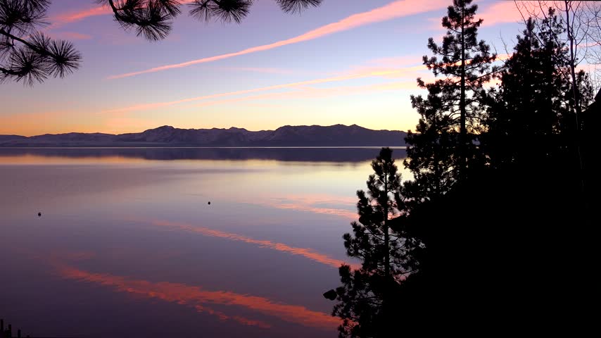 Breathtaking landscape view of tranquil lake at dusk featuring soft pink clouds reflected on water surface and majestic mountain peaks silhouettes.