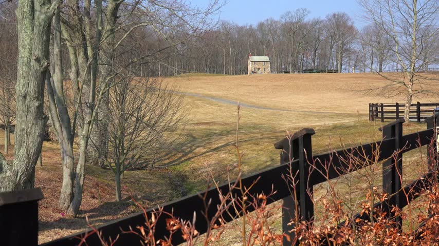 Stone building sits atop rural hill featuring dry grassy fields wooden fence foreground and leafless trees cinematic view.