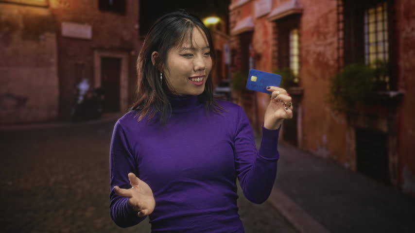 Woman holds blue creditcard in raised hand while gesturing with other hand on a narrow cobblestone street; surprise shopping.