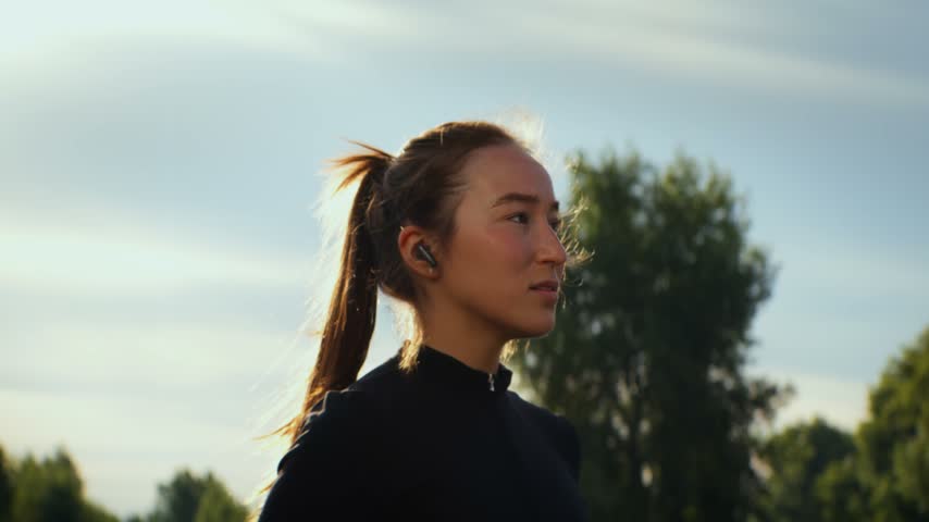 Young woman adjusting sunglasses and earbud outdoors in sunlit park with blurred trees and sky scene