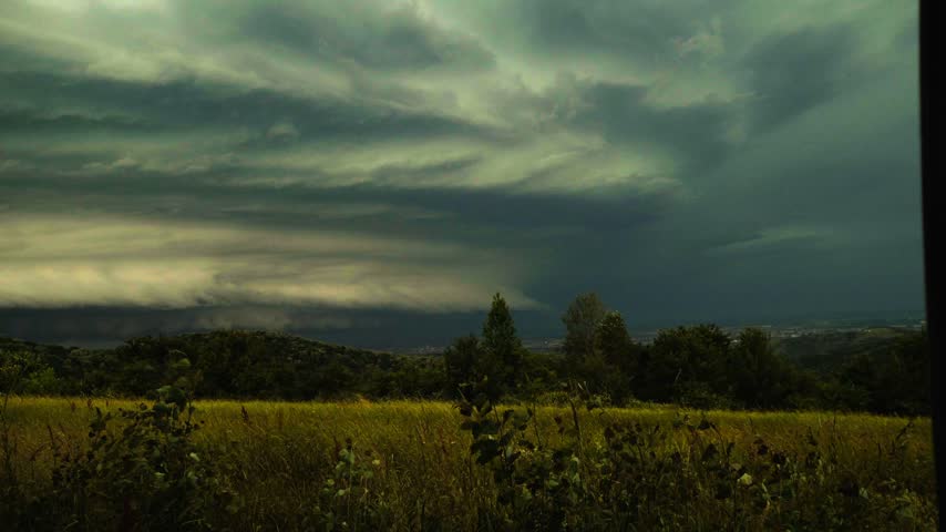 Dramatic dark storm clouds with bright lightning flashes rolling over a green rural landscape with trees and distant hills.