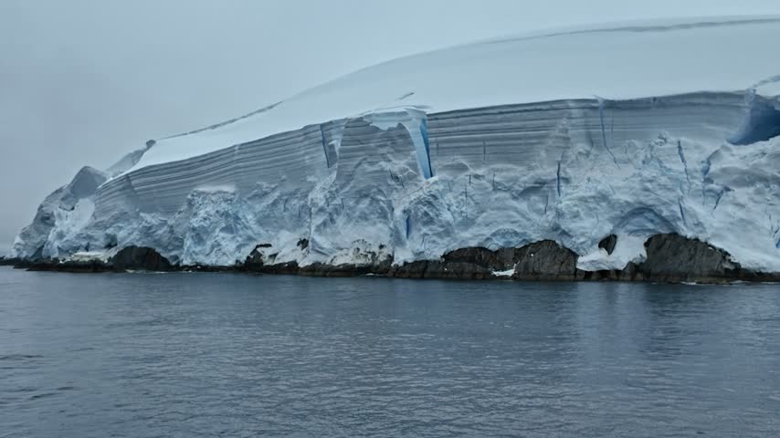 Blue-white ice cliffs with vertical breaks extend along a rocky shore above calm gray water in light rain.