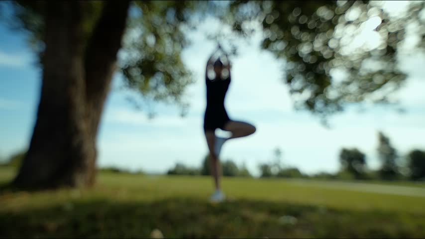 Blurred person holding tree pose with arms stretching in sunny park under large trees and open lawns
