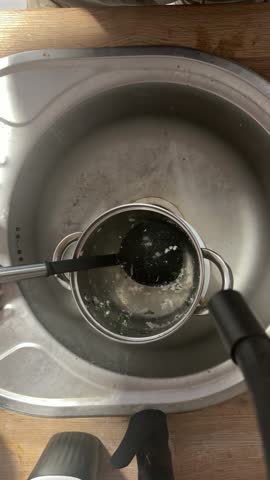 Top-down view of a metal saucepan with food residue and a black ladle resting inside a stainless steel kitchen sink.
