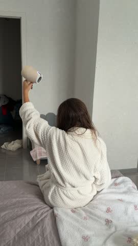 Seen from behind, a person in a white robe sits on a bed and aims a handheld hair dryer toward long dark hair.