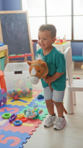 Boy holding a plush stick horse toy, smiling and rocking it near foam puzzle mat and toy cars in a daycare building; joy playfulness.
