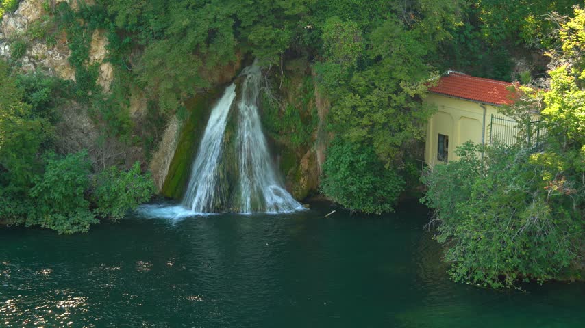 Beautiful waterfall flowing into a river next to a small house with a red roof surrounded by lush green trees