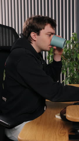 Consuming coffee, Young mug, Green stripes. Young man, dressed in black hoodie, drinks coffee from blue cup, with green plant and vertically striped wall serving as backdrop.