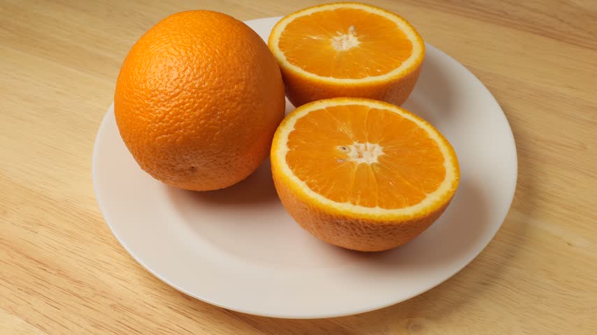 Freshly cut oranges and a whole fruit rotating on a white plate over a wooden table background