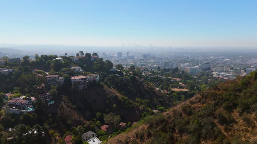 Aerial view over Hollywood Hills California. Luxury hillside homes with pools above steep canyon slopes. Hazy downtown Los Angeles skyline on sunny summer day. Peaceful real estate flight.
