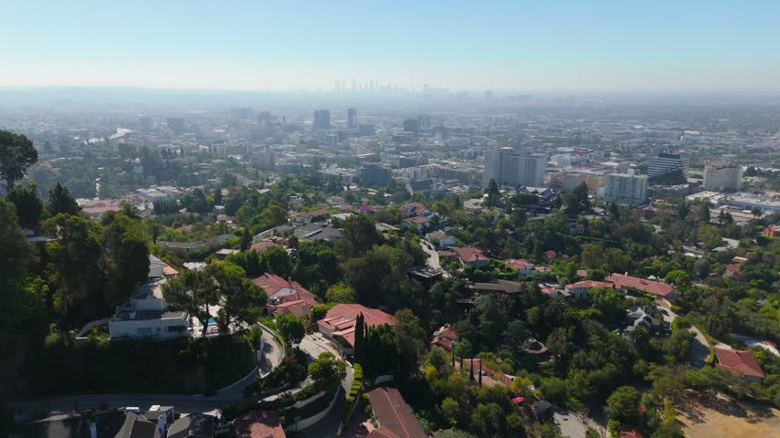 Hollywood Hills California aerial view over luxury hillside homes with pools and green trees. Downtown Los Angeles skyline in haze. Peaceful sunny day drone flight above famous real estate.