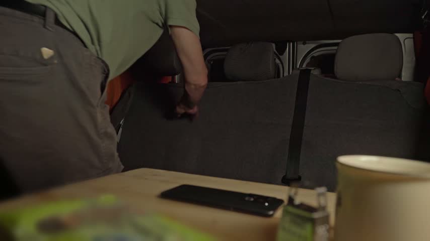 Man folding down vehicle rear seat to set up camper bed with pillows inside conversion van.