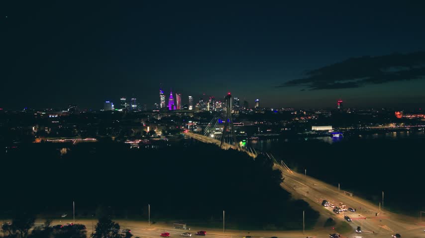 Stunning aerial footage capturing the vibrant Warsaw skyline at night, showing the illuminated downtown with its modern skyscrapers and the Świętokrzyski Bridge over the Vistula River