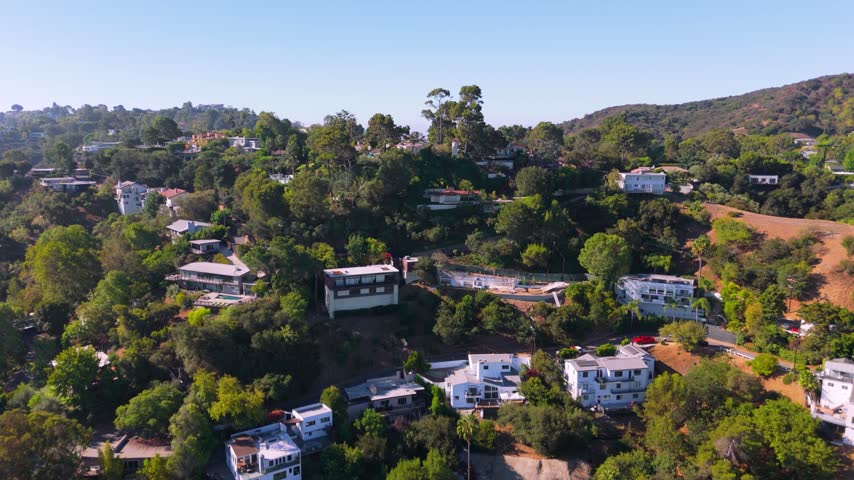 Aerial view over Hollywood Hills California. Luxury homes with pools hidden among green trees on canyon slopes. Los Angeles skyline in hazy distance. Peaceful sunny day in upscale real estate area.