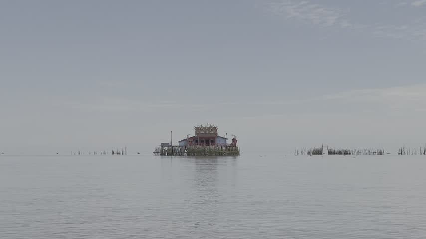A peaceful waterfront scene featuring a traditional Chinese temple standing on stilts over calm water, surrounded by blue skies and a serene natural landscape.