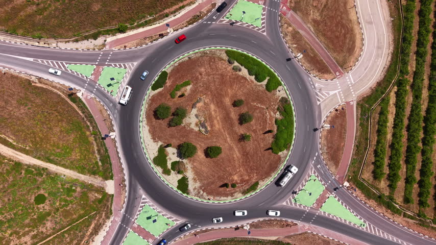 Overhead top-down view of a large circular roundabout with a landscaped central island featuring dry soil and green shrubs, surrounded by cars in traffic