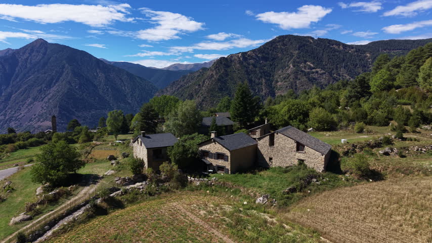 Rustic stone farmhouses of Engolasters hamlet nestled in a green alpine valley surrounded by forested Pyrenees mountain peaks under a blue sky with white clouds