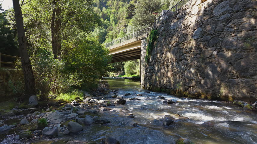 A concrete road bridge with iron railing spans a clear rocky mountain stream, flanked by a stone retaining wall covered in ivy, surrounded by lush green forest trees