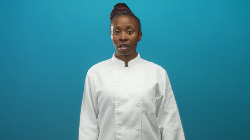 African american woman chef touching forehead with hand, wearing white chef jacket and bun against blue studio backdrop; stress.