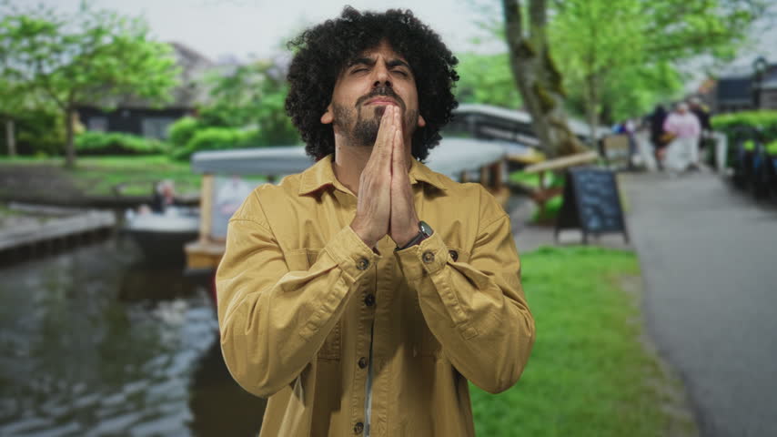 Man with curly hair and hands pressed together praying on a street by a canal in amsterdam wearing a mustard jacket; hope reflection.