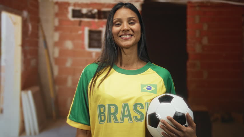 Woman holding soccer ball and smiling with eyes closed in brick building, wearing brazil jersey and t shirt, hand on ball; team pride.