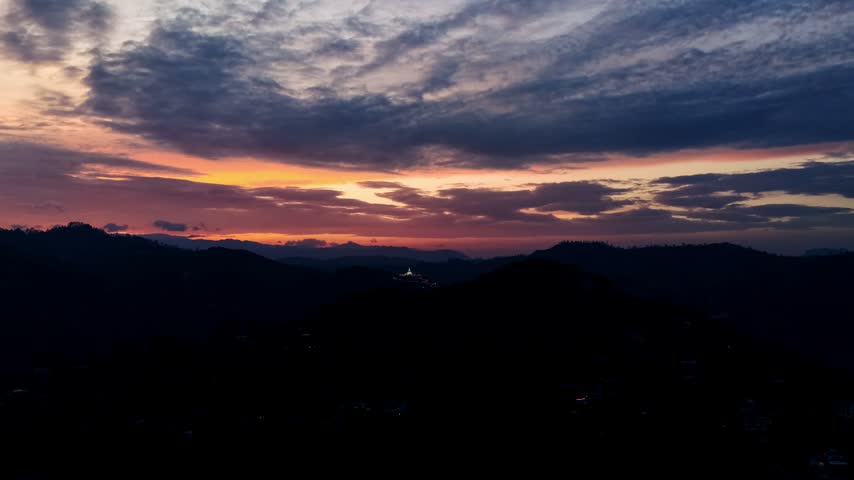 Dramatic sunset-to-blue-hour aerial timelapse over the hill country near Ella, Sri Lanka, with moving clouds, layered mountain silhouettes and the illuminated Mahamevnawa Buddhist Monastery and white stupa glowing in the distance.