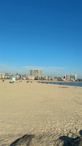 Vertical view across sandy Long Beach shoreline with foreground rocks, calm ocean, beachgoers and waterfront apartment buildings beneath a clear blue sky