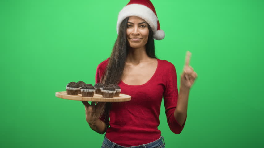 Woman holds wooden tray of chocolate muffins and shows peace sign with right hand in studio; holiday cheer sharing.