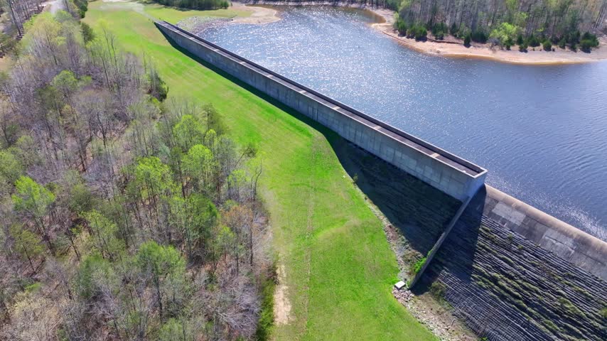 Panning aerial view of a concrete dam blocking shimmering deep blue water of a reservoir with grass cover banks on a sunny day,