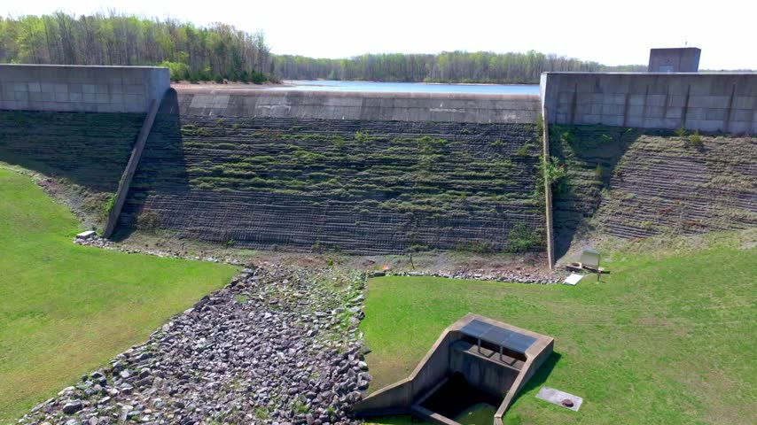 Rising aerial view of a concrete dam blocking shimmering deep blue water of a reservoir with tree lined sandy shores on a sunny day.