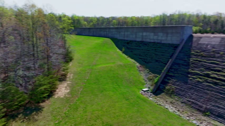 Low panning aerial shot of a concrete dam blocking shimmering deep blue water of a reservoir with tree lined sandy shores and grass-coverd banks on a sunny day,