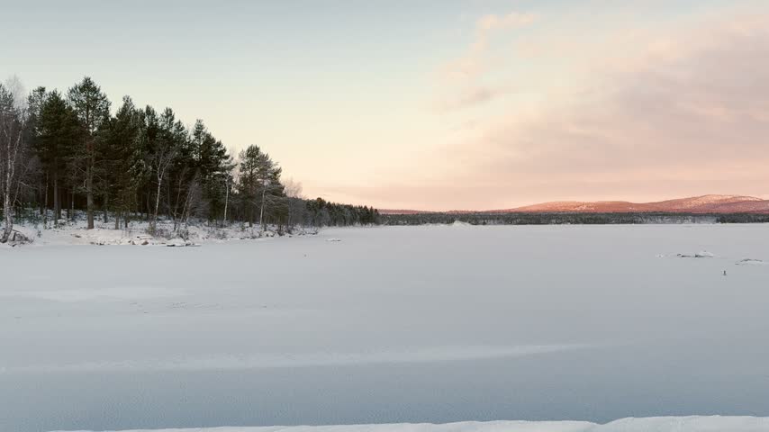 A quaint wooden house sits peacefully by the edge of a vast, snow-covered frozen lake, flanked by a sparse forest under a soft, clear winter sky in Finnish Lapland. Captures authentic Nordic winter living.