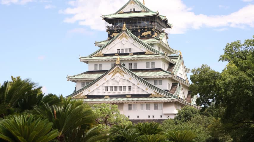 View of the iconic main tower of Osaka Castle in Japan, showcasing traditional Japanese castle architecture surrounded by greenery beneath a bright sky.