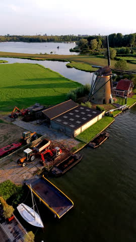 Vertical aerial drone video shot providing a high-angle view over an agricultural riverbank yard in Warmond, South Holland, Netherlands. The camera angle looks down at a traditional Dutch brick windmill standing beside a farm shed, with tractors, trailers, and excavators parked on the paved work area. Barges filled with soil sit moored along the riverbank, with a yellow utility boat and a small sailboat anchored in the foreground water. In the background, green polder meadows, winding drainage canals, and wide blue lakes extend toward distant treelines and the far city skyline under a warm, soft sunset sky during golden hour. High-resolution stock media highlighting Dutch rural farming, historic windmill heritage, inland waterway transport, polder agriculture, and scenic South Holland countryside landscapes. Ideal for vertical social media reels, mobile video production, travel stories, commercial advertising, and broadcast media.