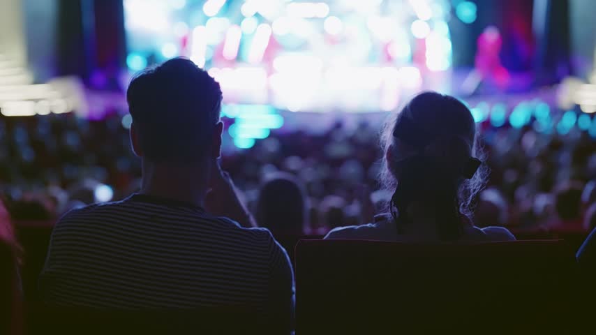 A couple sits in a darkened theater, their backs to the camera, intently watching a vibrant, blurred stage performance. The scene captures an authentic shared entertainment experience.
