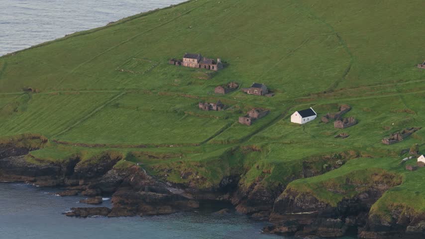 Aerial view of scattered white cottages on the green hillside of Great Blasket Island above rocky Atlantic cliffs.