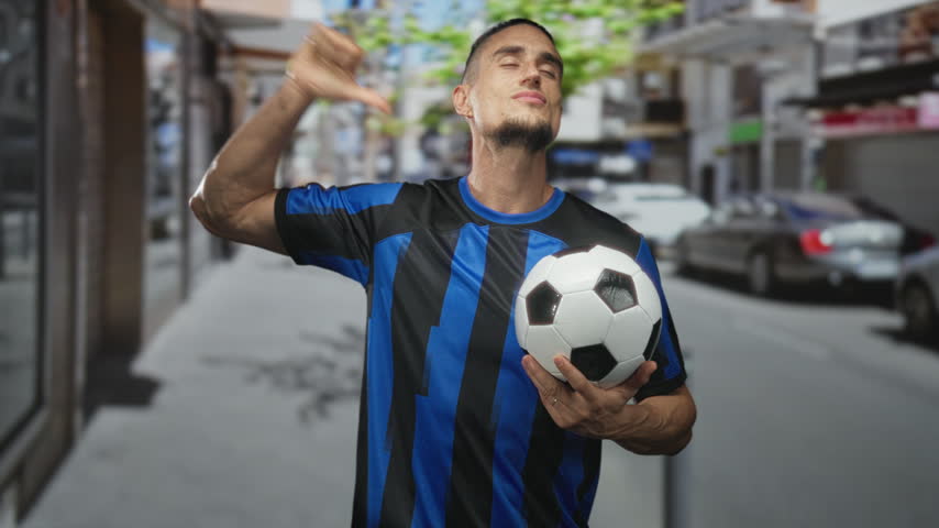 Man in blue and black striped soccer jersey holding a soccer ball in one hand and giving thumbs down with the other on a street with parked cars and storefronts; disappointment resilience.