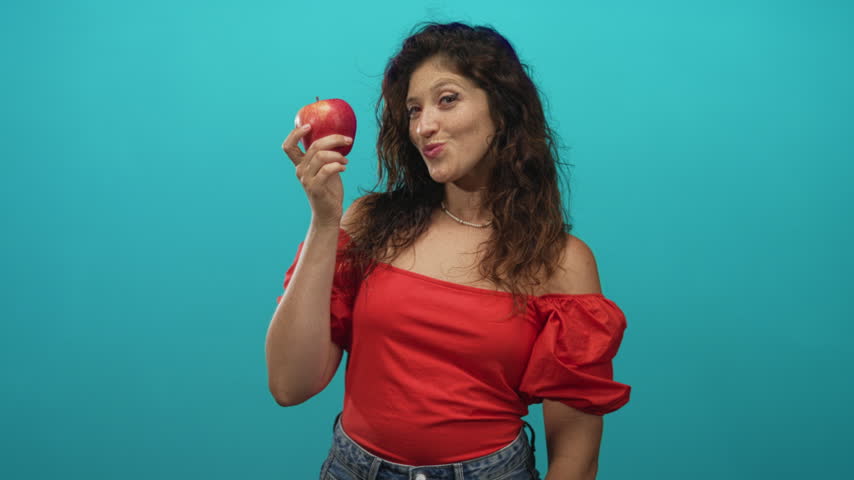 Woman holding red apple in studio turquoise, hand raised near face and smiling; playful healthy confidence.