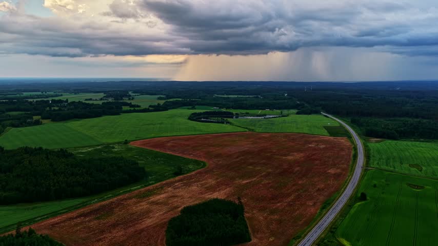 Heavy rain falling from dramatic storm clouds over green and brown agricultural fields and a highway