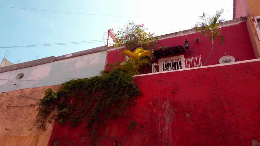 Panning across colorful residential buildings with red and orange stucco walls. Barred windows tiled roofing and tropical plants create a distinctive Mexican streetscape.