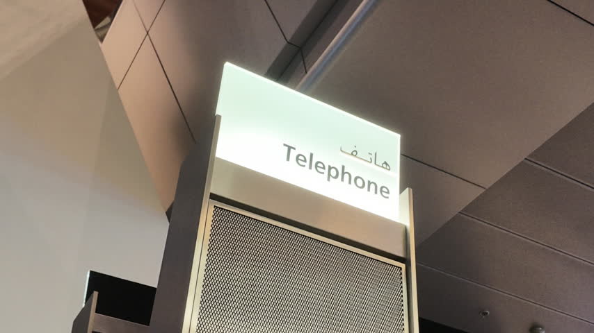 Illuminated public telephone sign featuring English and Arabic text inside a modern airport terminal. Static 4K slow motion shot of communication wayfinding signage.
