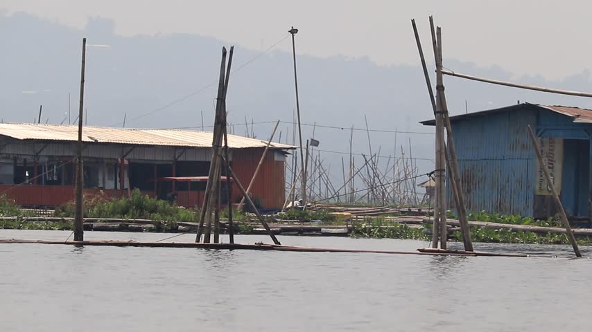 A view from the water showing rustic stilt houses and bamboo structures of a traditional floating fishing village under a cloudy sky