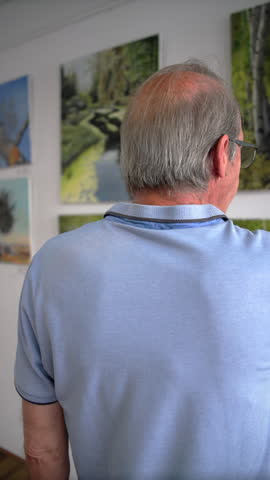 A senior man gestures toward a painting of birch trees while examining a wall of landscape artwork, Slow Motion.