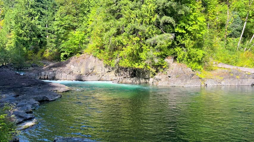 4K footage of turquoise whitewater flowing into a green river pool on the Campbell River in Elk Falls Provincial Park, Vancouver Island, British Columbia, Canada. Ripples and forest reflections fill the foreground beneath sunlit rock faces and overhanging evergreen trees.