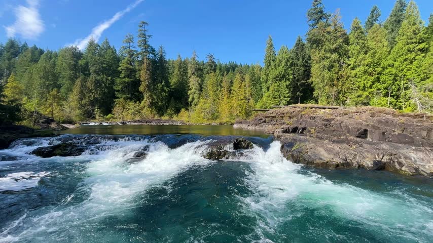 4K footage of Campbell River rapids spilling over low bedrock ledges into a swirling turquoise pool in Elk Falls Provincial Park, Vancouver Island, British Columbia, Canada. Foaming currents fill the foreground beneath sunlit rock shelves, evergreen forest and blue sky.