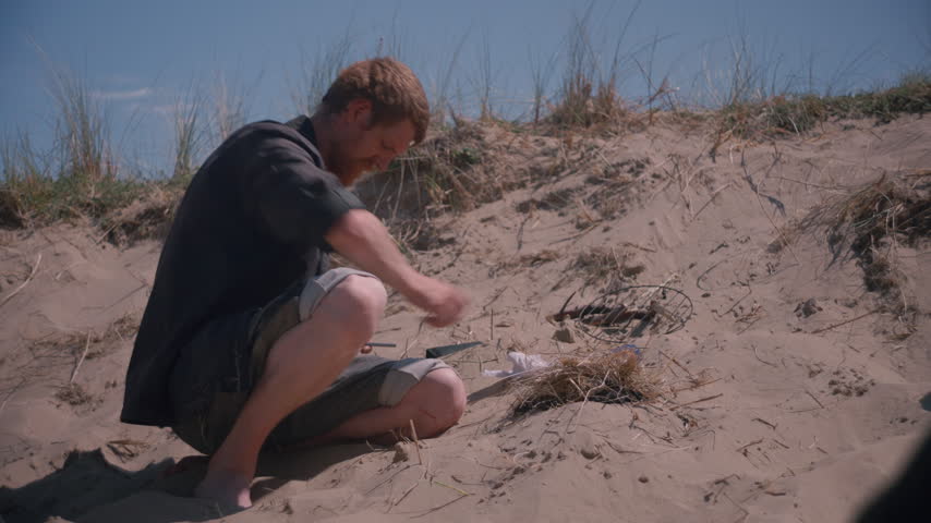 A man kneels by a small fire, blowing gently on the embers and sticks to help the flames catch.