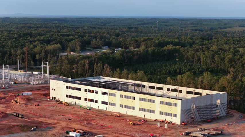 Long two story data hall in cream painted precast panels in Louisa County, Virginia, with spare wall panels stacked at the gable end and the substation beyond the pines. Aerial wide shot