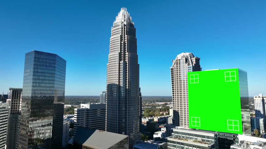 A tracked green screen billboard hangs among the uptown towers of Charlotte, North Carolina, corner markers on the panel and the Bank of America Corporate Center behind. Aerial lateral wide shot