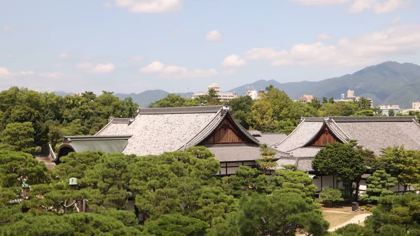 Elevated view across the traditional rooftops of Ninomaru Palace at Nijo Castle in Kyoto, Japan, with surrounding greenery, city scenery and distant mountains